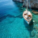 A sailboat with wooden trim, available for bareboat charters, is anchored in clear turquoise water near a rocky shoreline. The water is so clear that the seabed and rocks are visible beneath the surface. The scene is peaceful and sunlit, suggesting a relaxing coastal location.