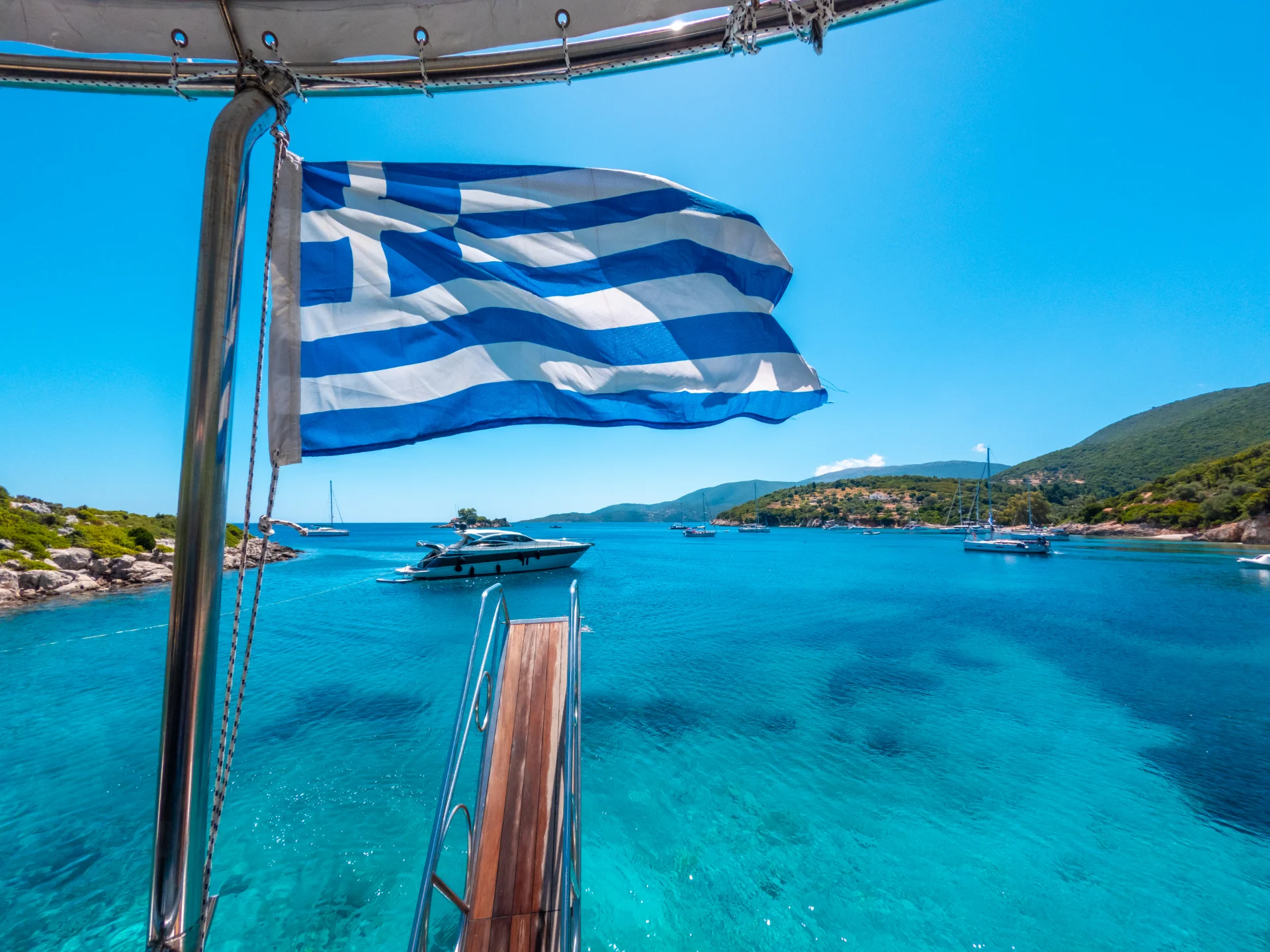 A Greek flag flutters in the breeze on a boat, overlooking a pristine turquoise bay with several sailboats and yachts. The coastline is dotted with green hills under a clear blue sky, evoking a serene Mediterranean scene.