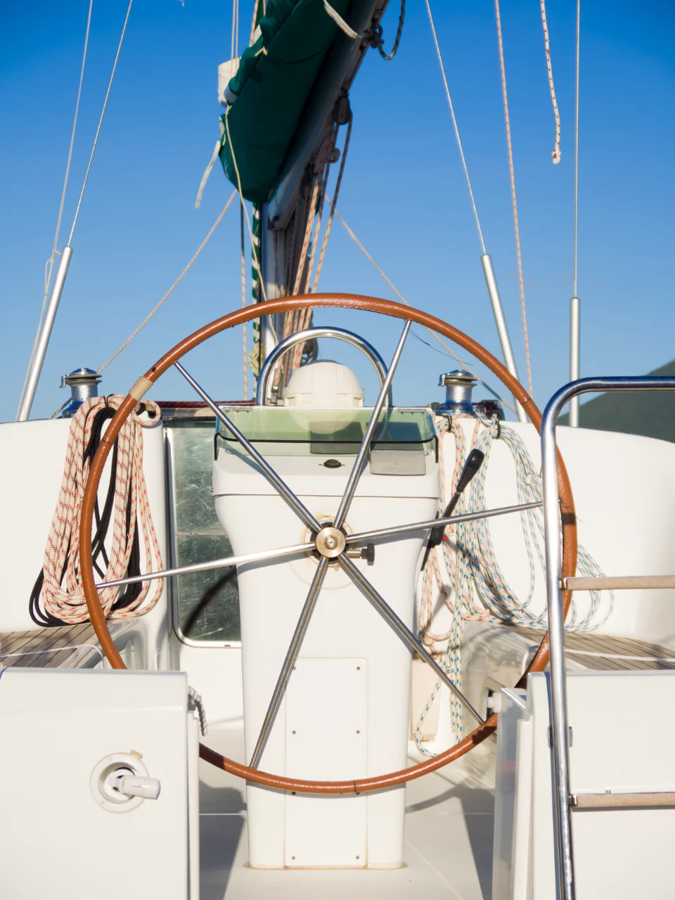 The photo shows the helm of a sailing yacht. It features a large wooden steering wheel at the center, surrounded by various ropes and seating areas on either side. The background displays a clear blue sky, contributing to the serene appearance.