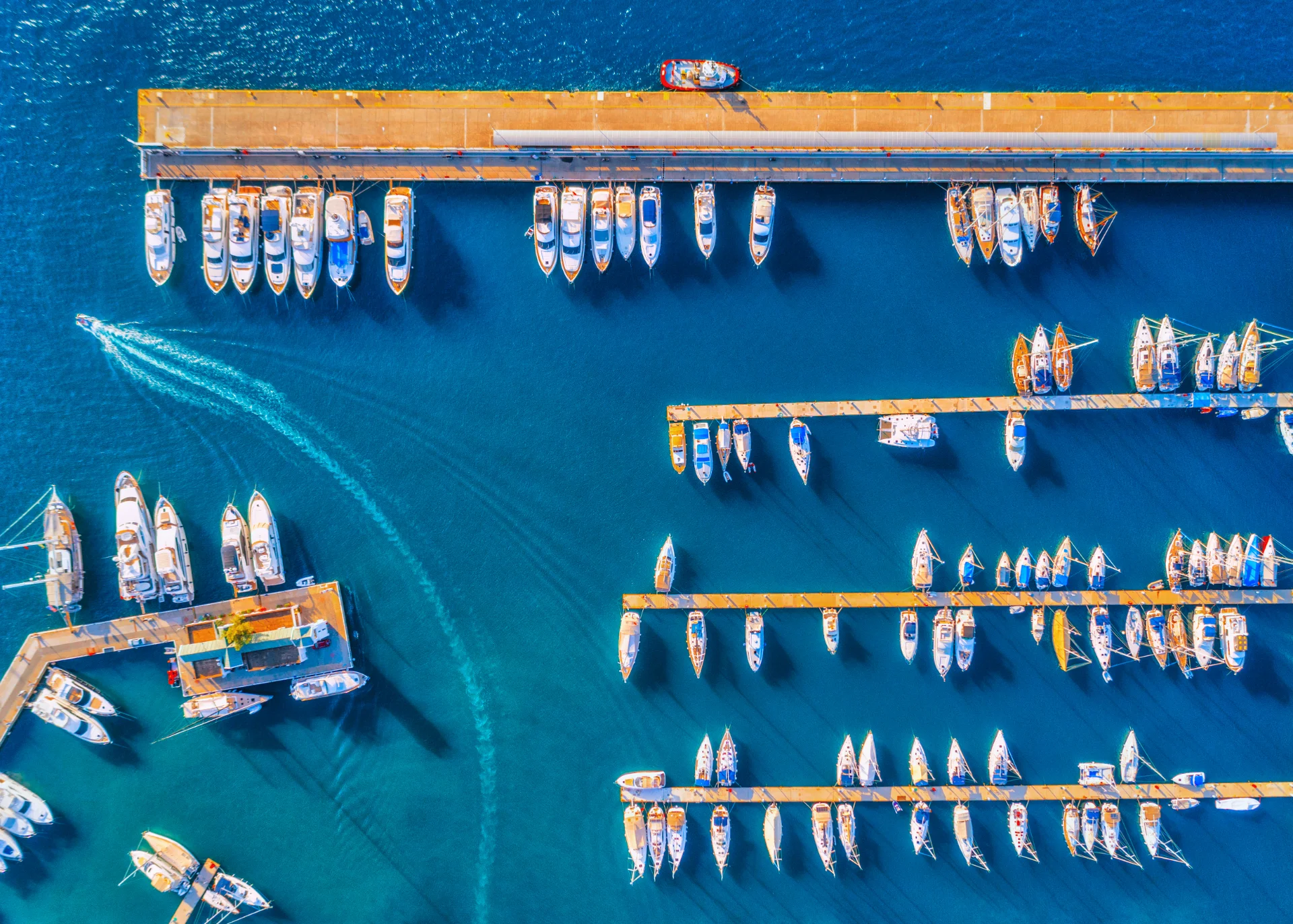Aerial view of a vibrant marina with multiple docks extending into deep blue waters. Numerous boats and yachts are moored alongside the docks, and a small boat leaves a curved wake as it moves away from the marina. The sunlight creates vivid reflections on the water.