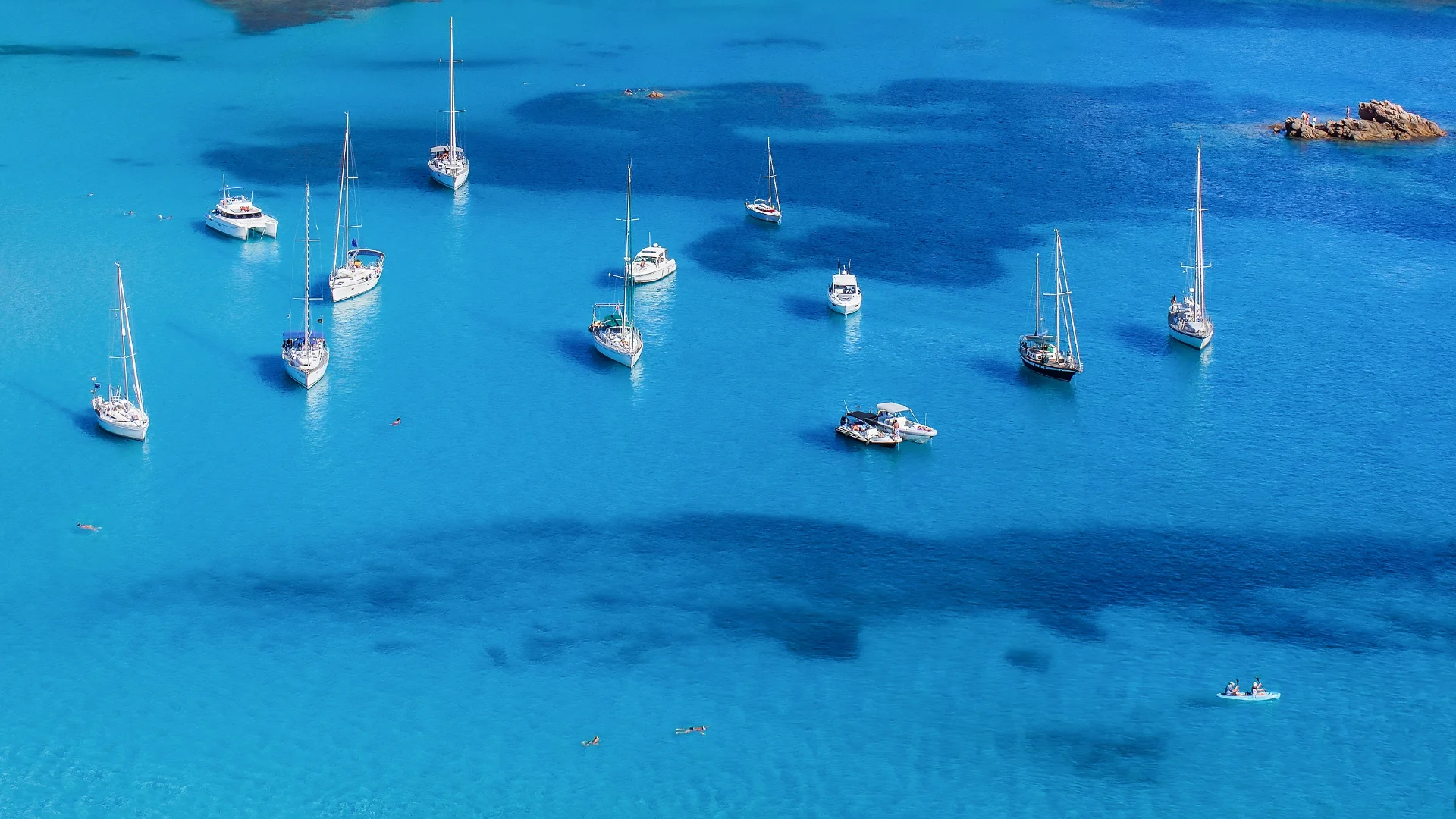 Aerial view of a serene turquoise blue ocean with several sailboats and yachts anchored, casting gentle shadows on the water. The image exudes tranquility with vibrant, clear water and a few swimmers visible near the boats.