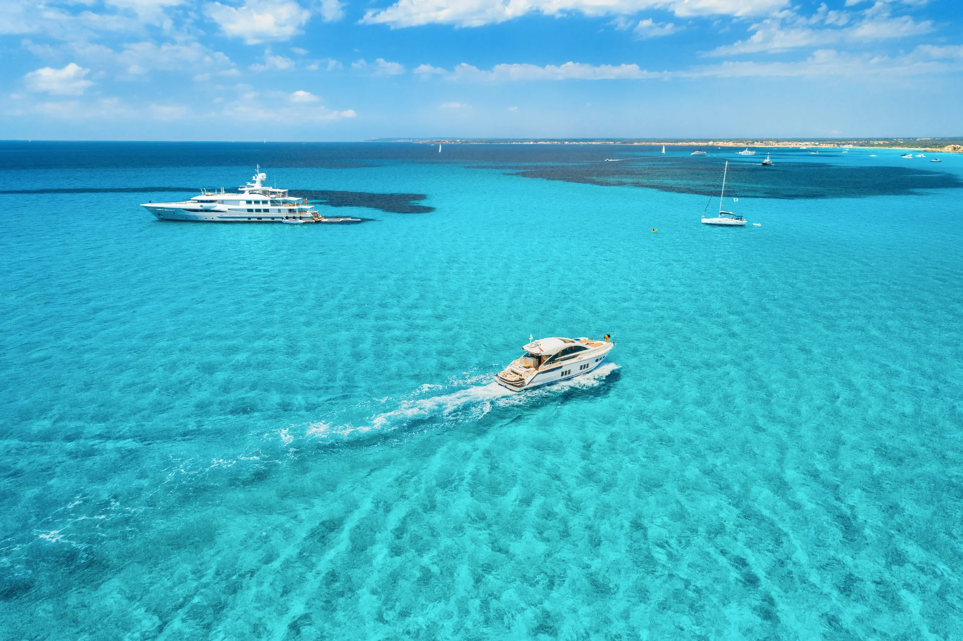 Aerial view of three yachts sailing through the crystal-clear turquoise waters of an ocean. The sky above is filled with scattered white clouds, and the horizon shows a strip of sandy shoreline with a few scattered buildings in the distance.