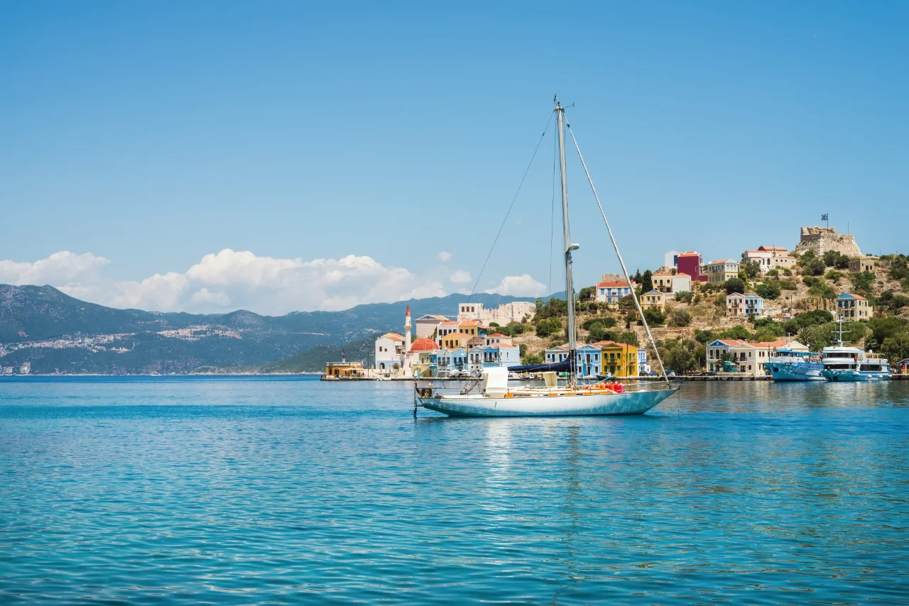 A sailboat is anchored on a calm, turquoise sea near a picturesque coastal village with colorful houses. The village is built on a hill, and lush mountains are visible in the background under a clear, blue sky.