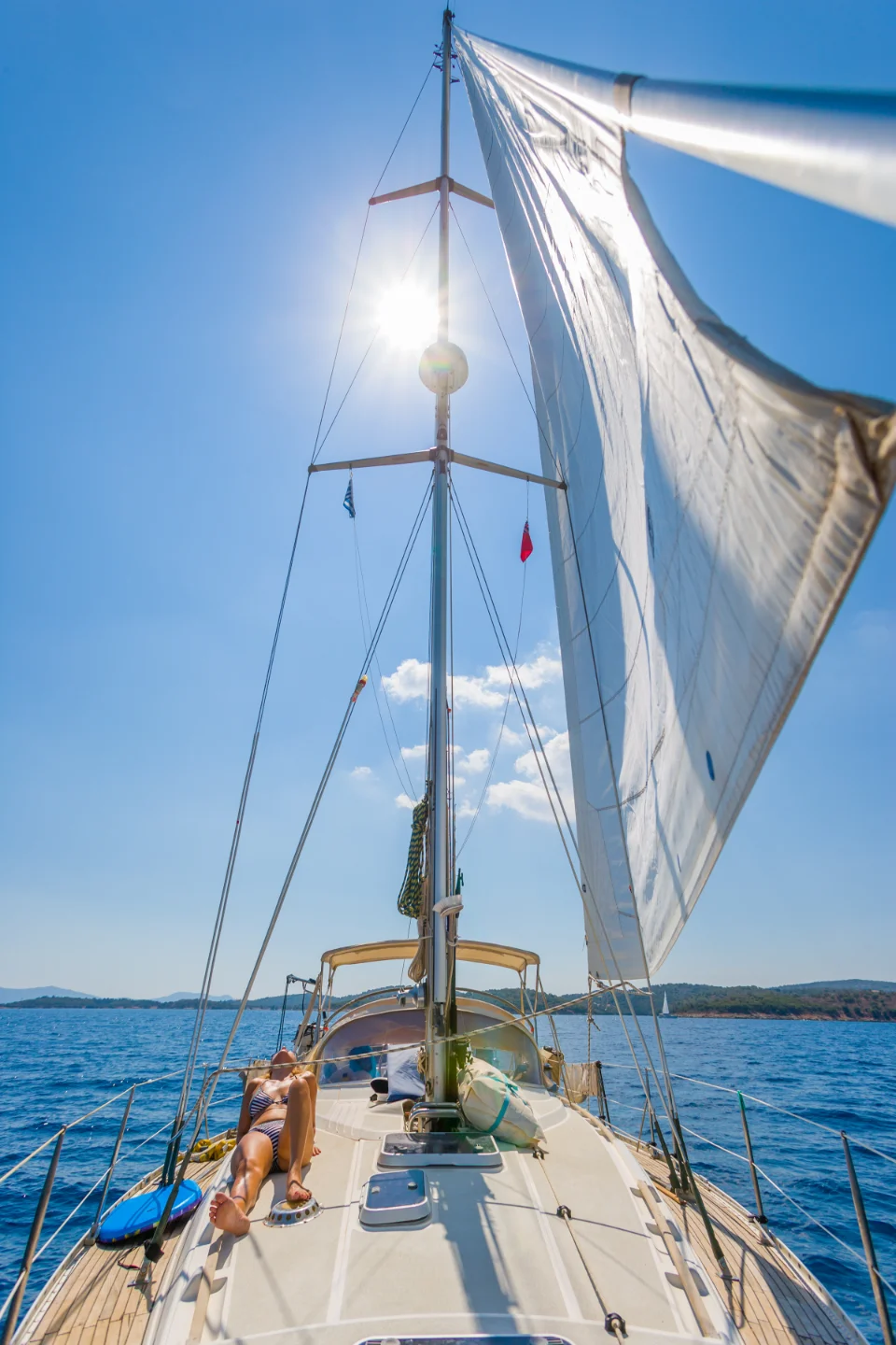 A person is lying on the deck of a sailboat under a sunny, clear blue sky. The sails are fully extended, and the boat is cruising on calm, blue waters. The sun is shining brightly, casting shadows on the deck, with distant land visible on the horizon.