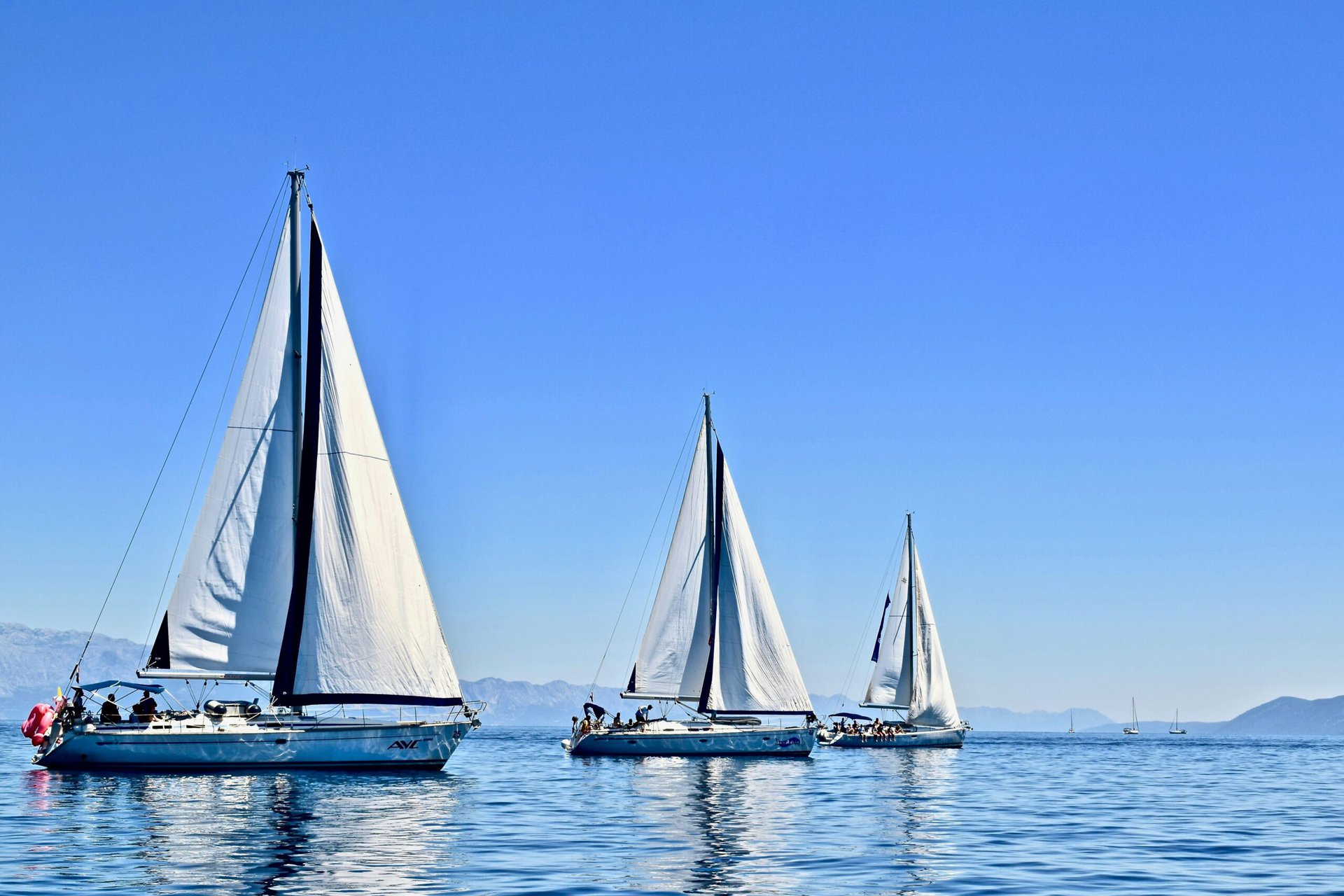 Three sailboats with white sails glide on a calm, blue sea under a clear sky. In the distance, mountainous terrain is faintly visible. The water reflects the sailboats, hinting at the serene and peaceful conditions of the scene.