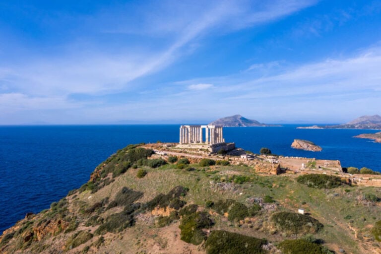 Aerial view of the ancient Temple of Poseidon at Cape Sounion, Greece. The site is perched on a cliff overlooking the vibrant blue Aegean Sea, with green, rocky terrain surrounding the ruins, under a clear blue sky. Islands can be seen in the distance.