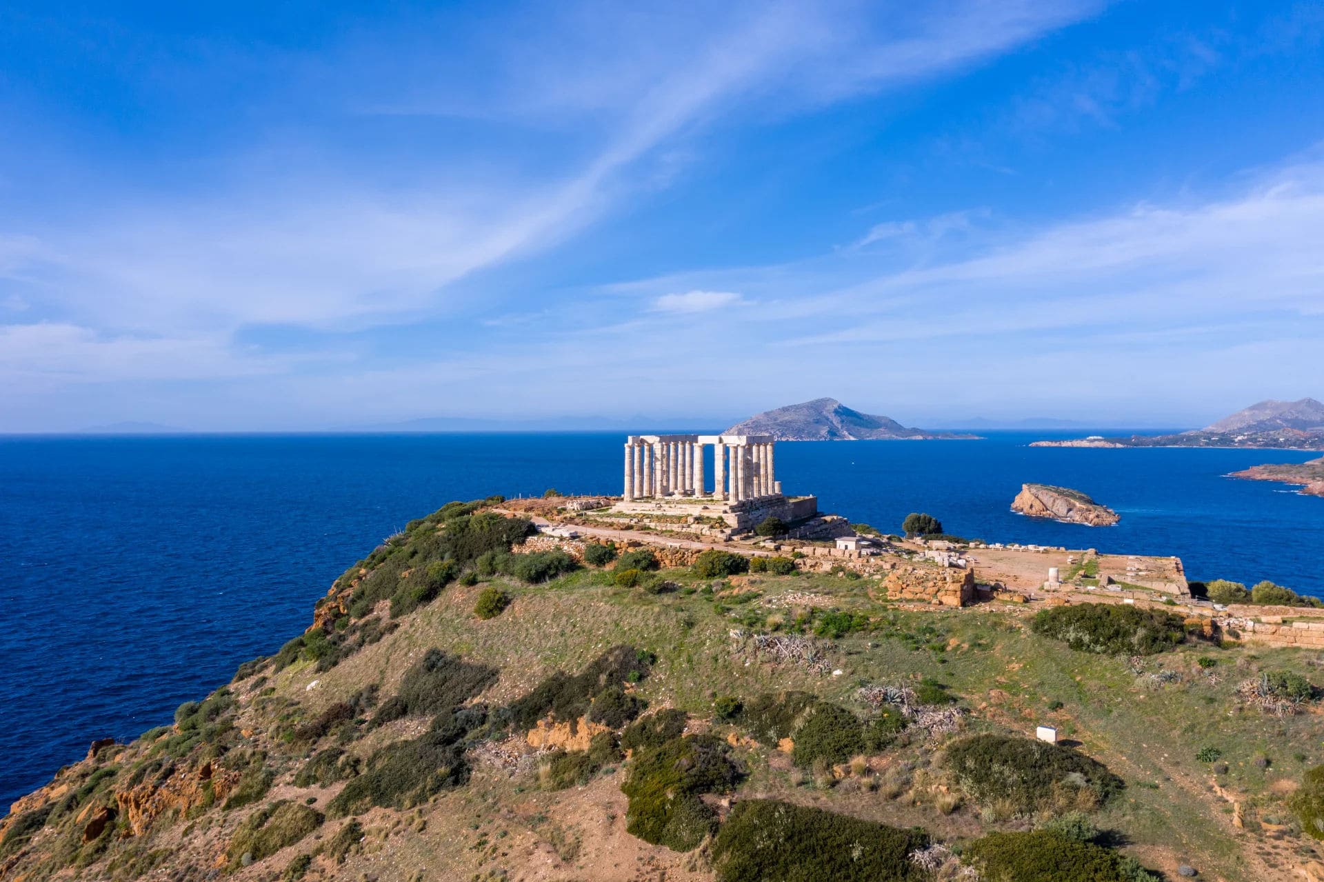 Aerial view of the ancient Temple of Poseidon located on a hill at Cape Sounion, Greece. The temple is surrounded by the bright blue Aegean Sea, with rocky islets and distant land masses under a clear, blue sky. The landscape is dotted with green shrubs and rocky paths.