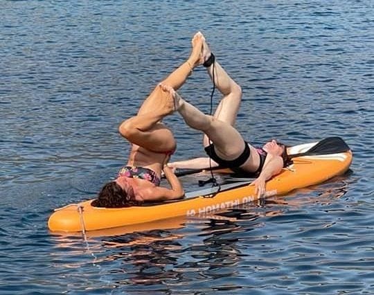 Two people are lying on their backs on an orange paddleboard in the water, performing a synchronized balancing pose with their intertwined legs raised in the air. Both are in swimwear and appear to be enjoying a sunny, clear day on the water.