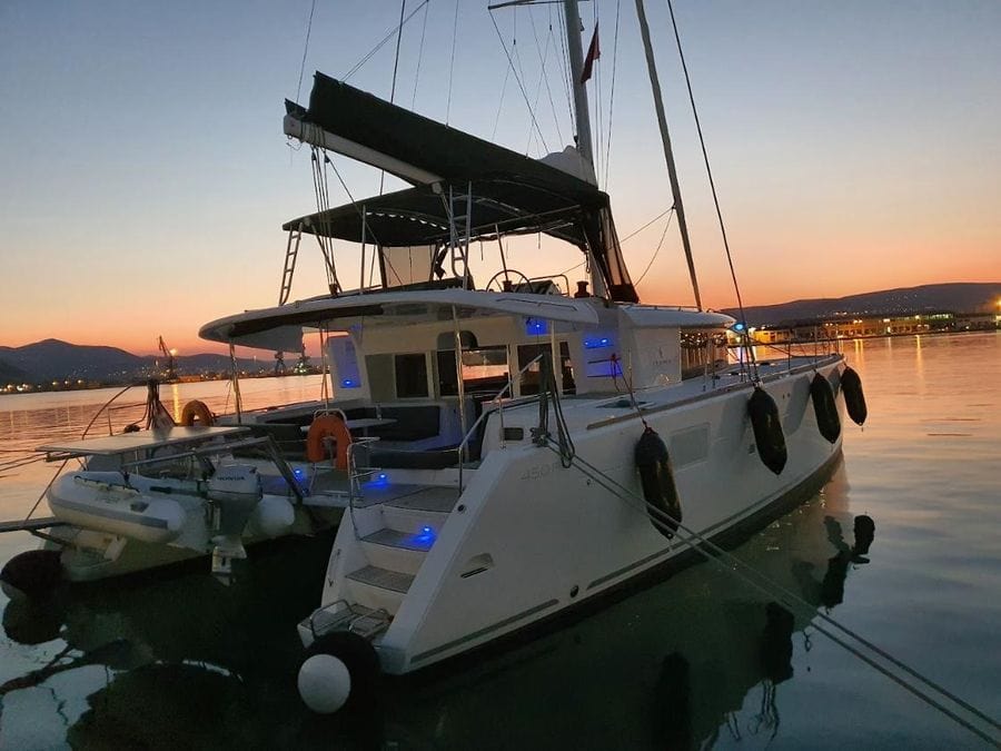 A luxurious sailboat docked at a marina during sunset. The boat is illuminated with blue lights and features a dinghy attached to its rear. Calm water reflects the boat and the colorful sky, with distant lights and silhouettes of mountains in the background.
