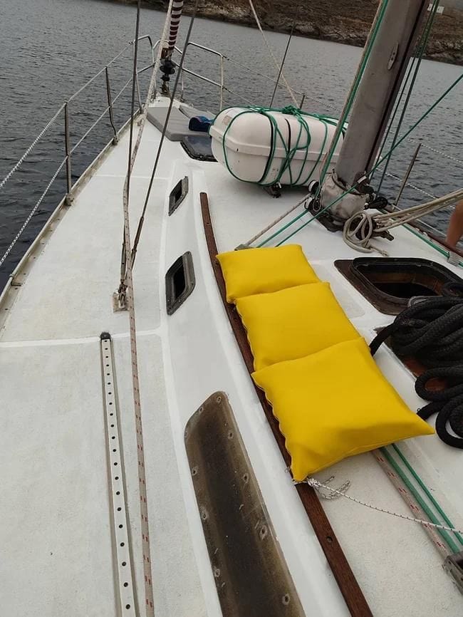 A perspective view of a sailboat's deck with two bright yellow cushions on the left side for seating. The mast and various rigging are also visible, along with a lifebuoy. The boat is docked in a calm, rocky coastal area.