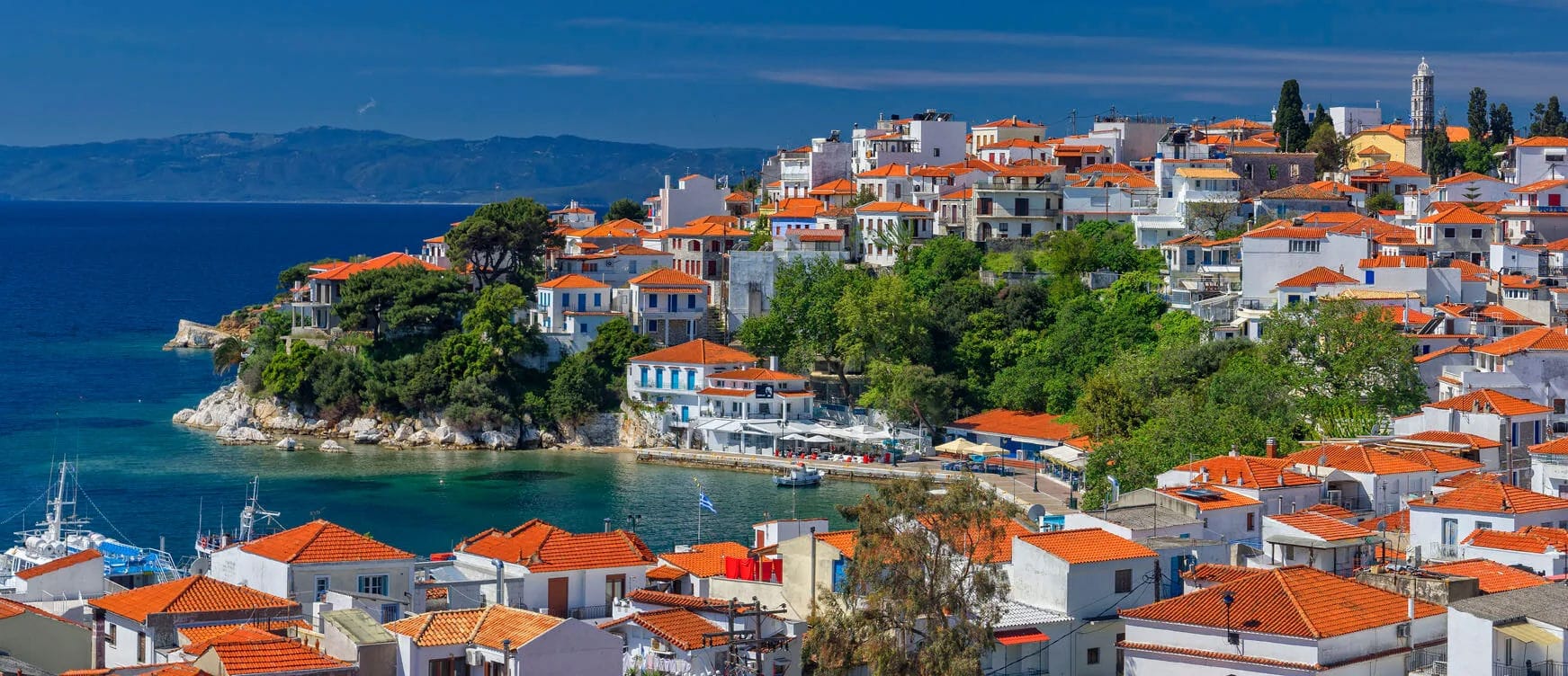 Scenic view of a coastal town with white buildings and red-tiled roofs under a clear blue sky. The town is nestled along a shoreline with boats docked in a small harbor. Green trees and hills surround the area, and distant mountains are visible across the water.