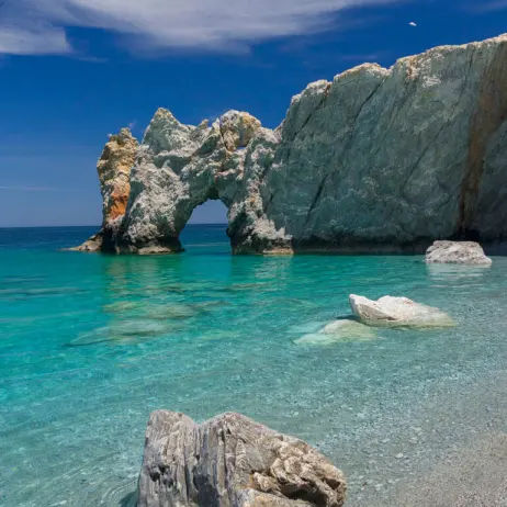 A picturesque beach scene with crystal-clear turquoise water, rocky formations, and a large, natural archway formed by rocks. The sky is a vibrant blue with a few wispy clouds. Several white rocks are scattered near the shoreline.