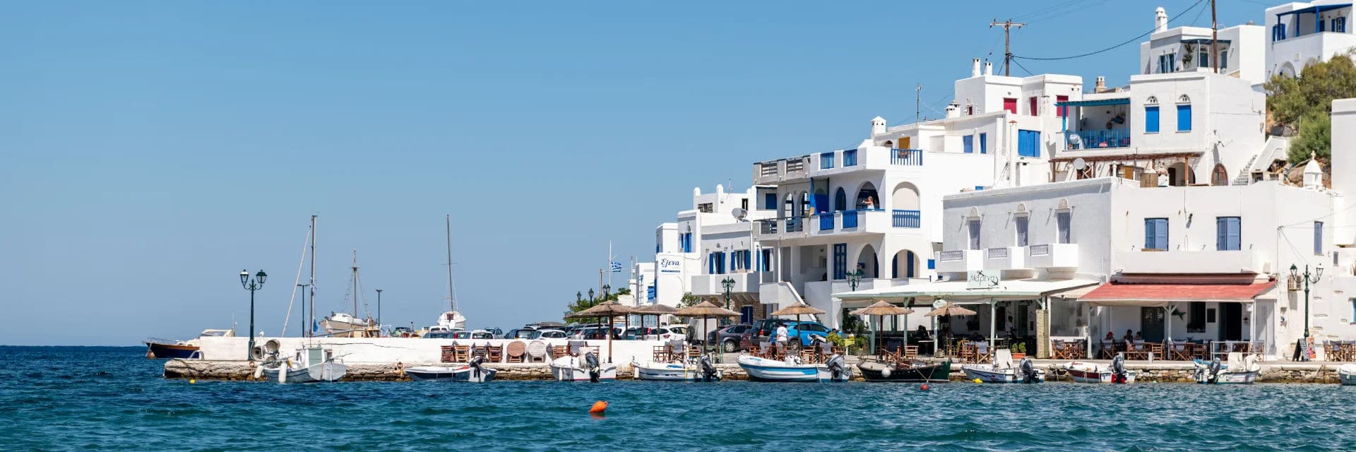 A scenic seaside view captures whitewashed buildings with blue accents along a waterfront. Several boats are moored at the dock, and a few outdoor cafes or restaurants line the shore, with patrons seated under umbrellas. The sky is clear and blue.

.